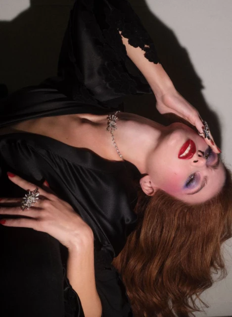 A woman with red hair and creative makeup, wearing jewelry and a black silk robe, poses against a neutral background in a studio setting.