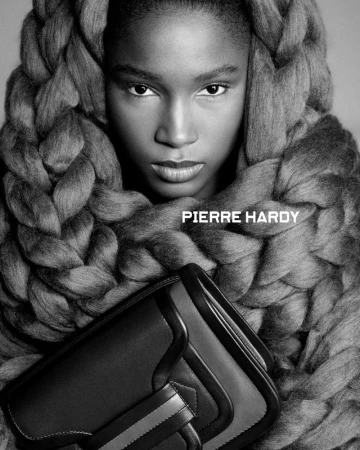 Close-up portrait of a Black woman framed by a large chunky knit braid, holding a structured dark leather handbag.