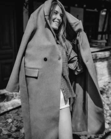 A smiling woman wearing an oversized wool coat draped over her shoulders outdoors in a black and white photograph.