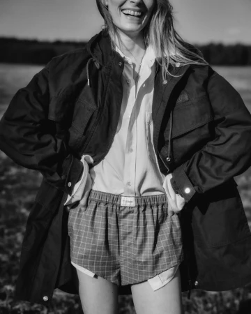 Monochrome fashion campaign image featuring a smiling model wearing a dark jacket, button-down shirt, and plaid shorts outdoors.
