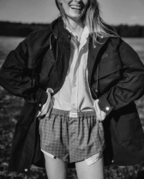 Monochrome fashion campaign image featuring a smiling model wearing a dark jacket, button-down shirt, and plaid shorts outdoors.