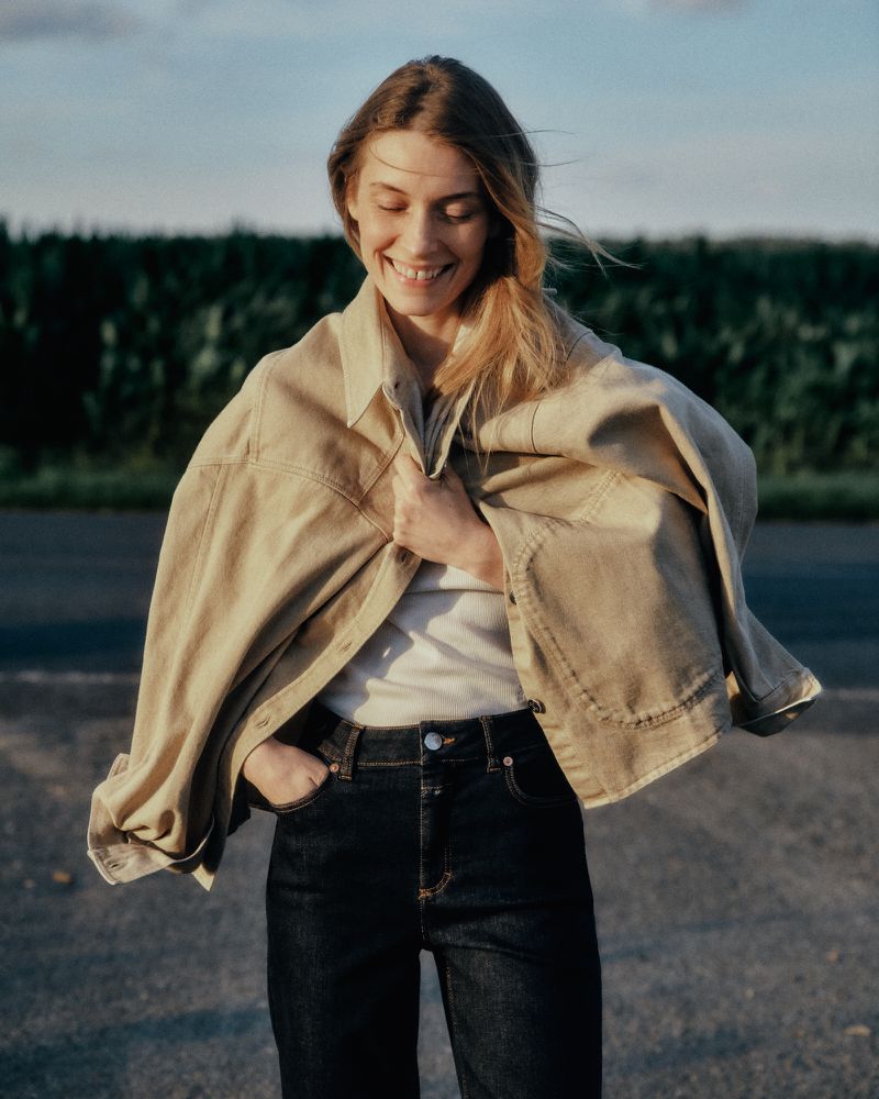 Smiling female model wearing a tan jacket draped over her shoulders with dark jeans against an outdoor backdrop