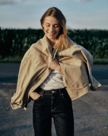 Smiling female model wearing a tan jacket draped over her shoulders with dark jeans against an outdoor backdrop