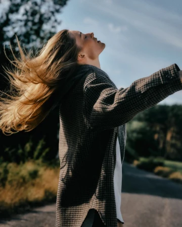Woman with long blonde hair whipping in the wind, wearing a patterned coat outdoors under an open sky