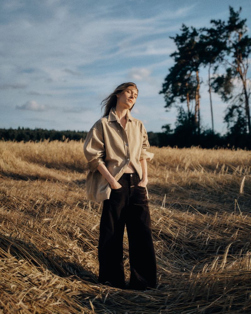 Model standing in a golden wheat field wearing a beige button-down shirt and dark trousers under a cloudy sky.