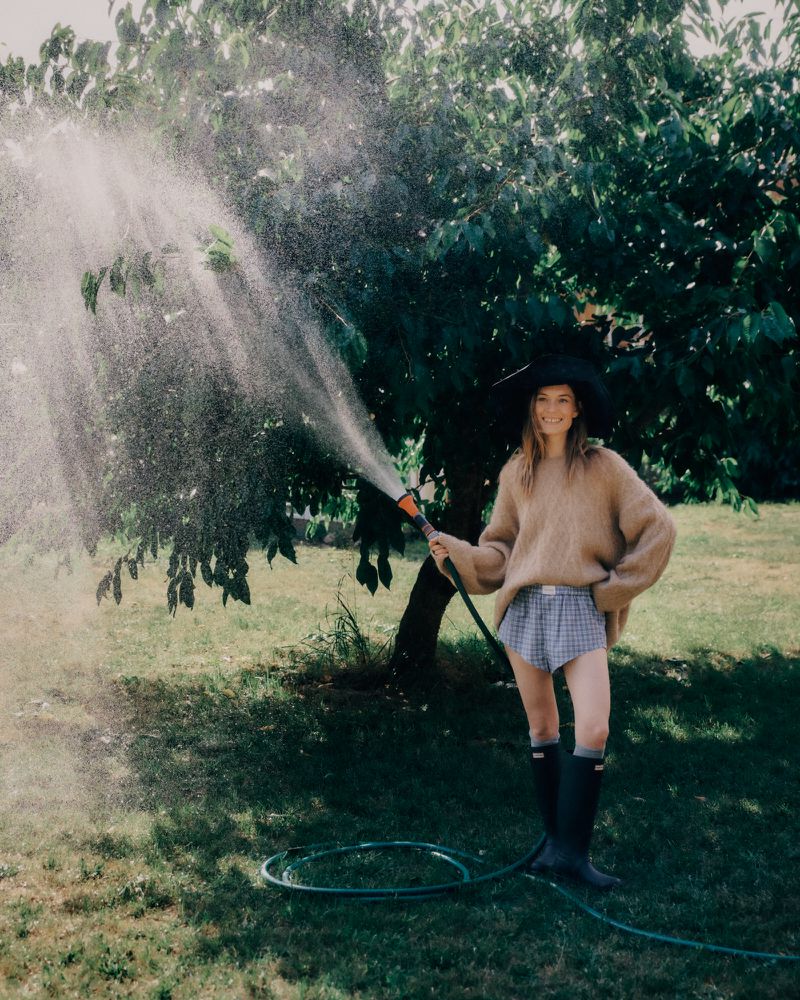 Model holding a garden hose spraying water in a lush green backyard for the Closed autumn winter campaign.
