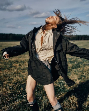 Model in dark jacket and light shirt tossing head back with hair flying in an open field under a blue sky.