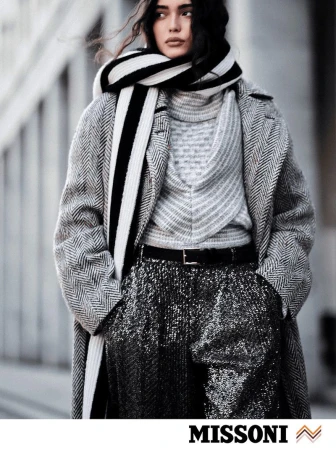 Model wearing a grey herringbone coat, sequin trousers, and a black and white striped scarf against an urban building exterior background.