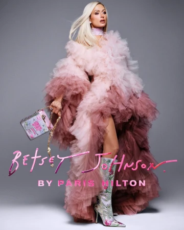 Paris Hilton in a voluminous pink tulle ruffled coat and heeled boots holding a handbag against a grey backdrop