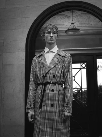 Monochrome portrait of male model Ethan Harwood in a plaid trench coat standing against an architectural backdrop.