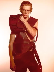 Portrait of a male model wearing structured dark apparel and holding a burgundy messenger bag against a warm gradient background