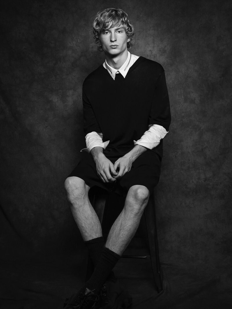 Portrait of male model Ethan Harwood seated on a stool wearing a dark layered outfit against a textured grey background.