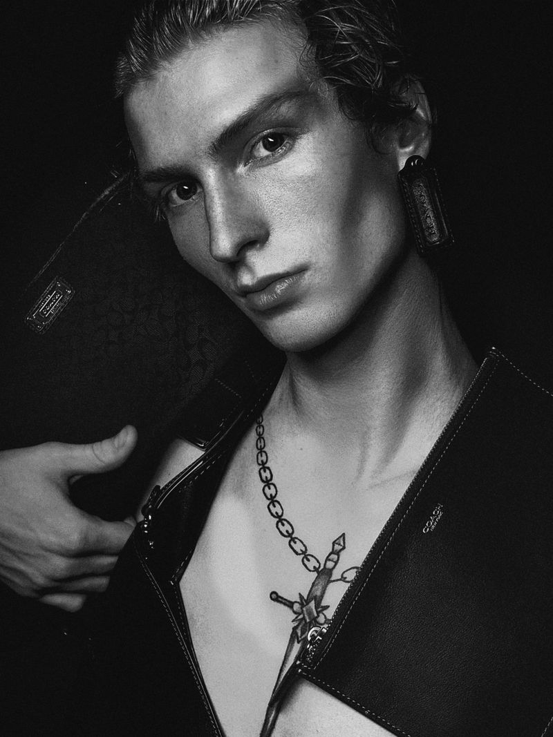Close-up monochrome portrait of male model Ethan Harwood wearing a dark structured vest and statement earring against a black background.