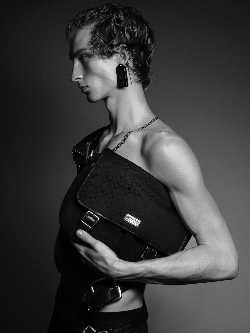 Monochrome portrait of male model Ethan Harwood holding a black messenger bag across his chest against a plain dark studio background.