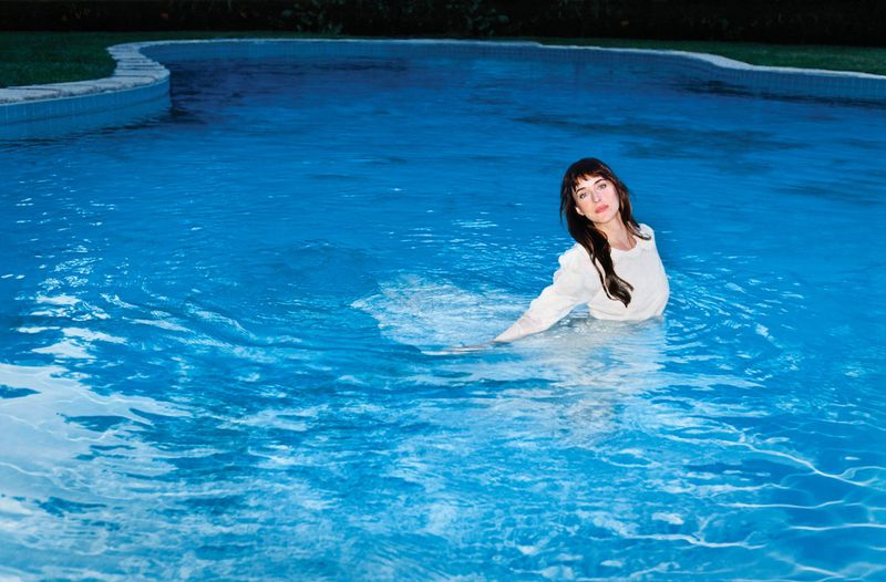 Dakota Johnson in a white top standing in a blue swimming pool for an American Vogue editorial portrait.