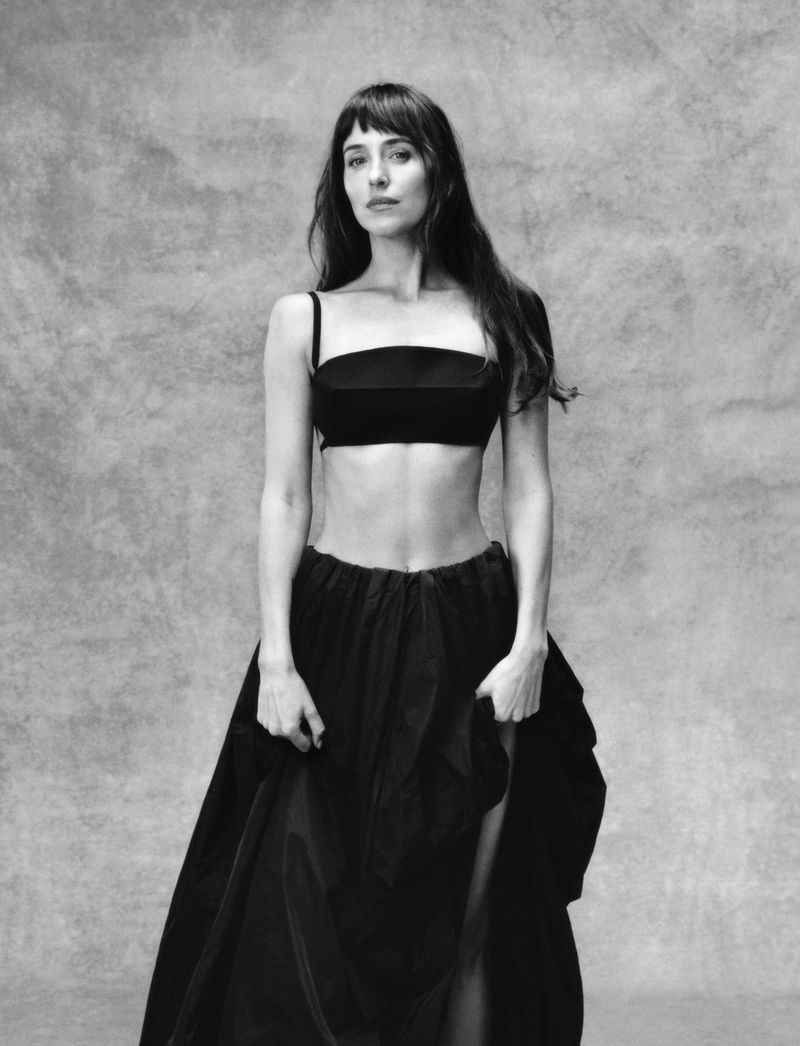 Dakota Johnson in a black two-piece outfit standing against a textured grey studio backdrop.