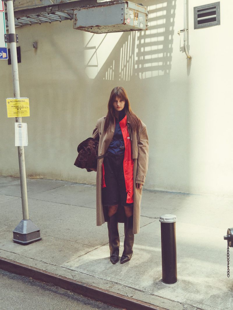Model standing on a city sidewalk wearing a beige trench coat, dark boots, and holding a patterned bag against a light wall
