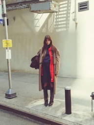 Model standing on a city sidewalk wearing a beige trench coat, dark boots, and holding a patterned bag against a light wall