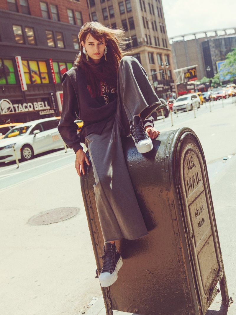 Model perched atop a large green U.S. mail collection box on a New York City street, wearing a dark sweatshirt and wide-leg trousers