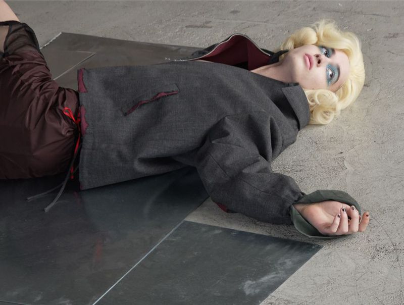 Blonde model lying on reflective metal panels wearing a dark grey jacket and brown trousers for AnOther Magazine editorial