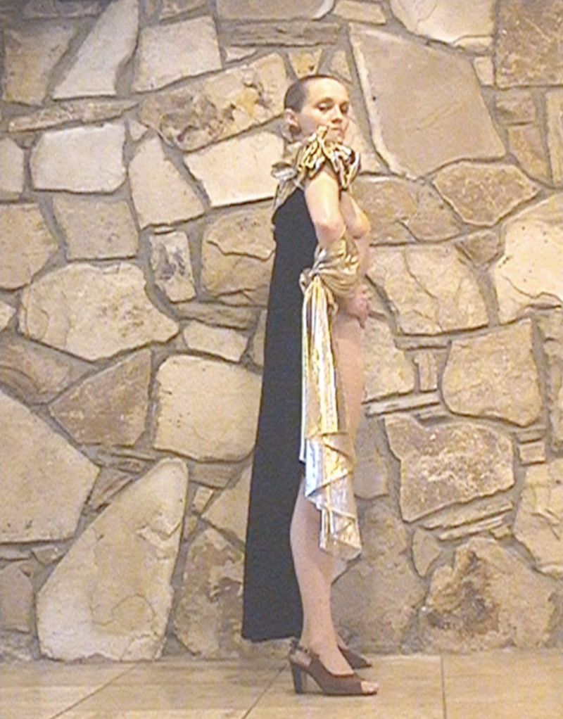 Woman poses in draped fabric gown against stone wall. She wears heels and elegant gown.  Neutral background, brown tones.
