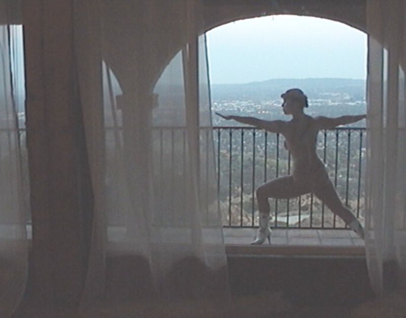 Adult female strikes a yoga pose in a sunlit room with white curtains, gazing out an arched window toward a cityscape and hills.