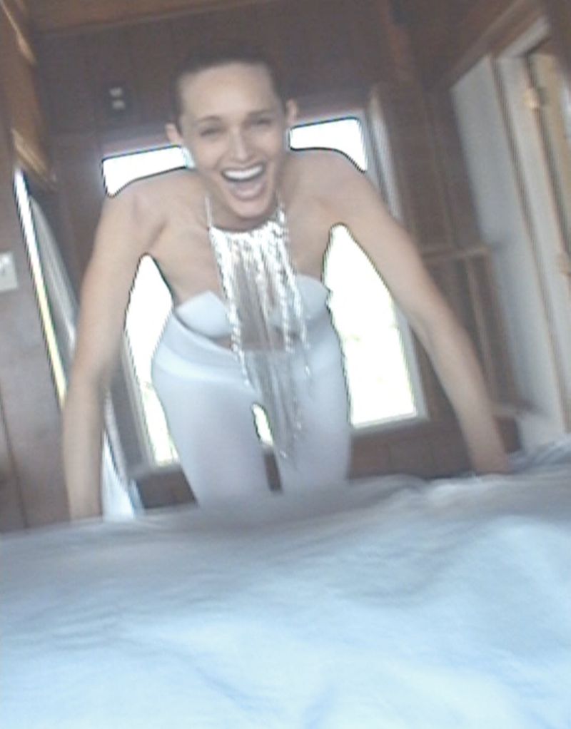 Smiling adult woman in white outfit and statement silver necklace posing indoors near a window with natural light, medium shot.