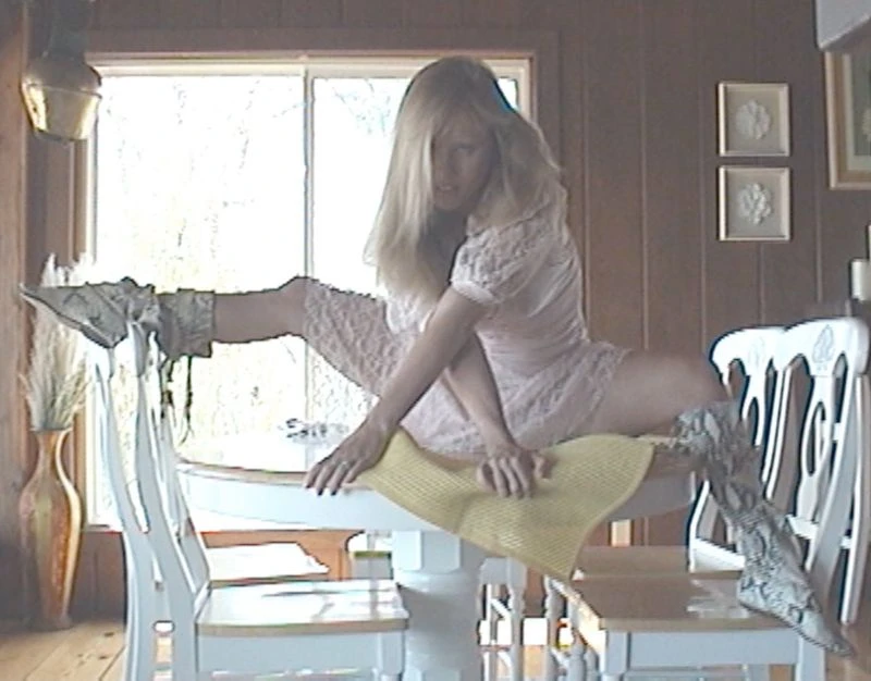 Female adult in pink dress and patterned high heel boots performs a split on top of a white table and chairs indoors near a window.