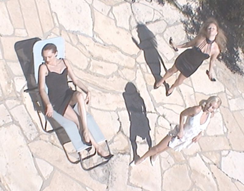 Overhead shot of three women in dresses, one reclining on a lounge chair, on a stone patio bathed in bright sunlight, casting shadows.