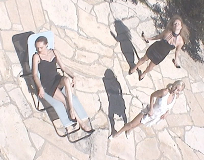 Overhead shot of three women in dresses, one reclining on a lounge chair, on a stone patio bathed in bright sunlight, casting shadows.