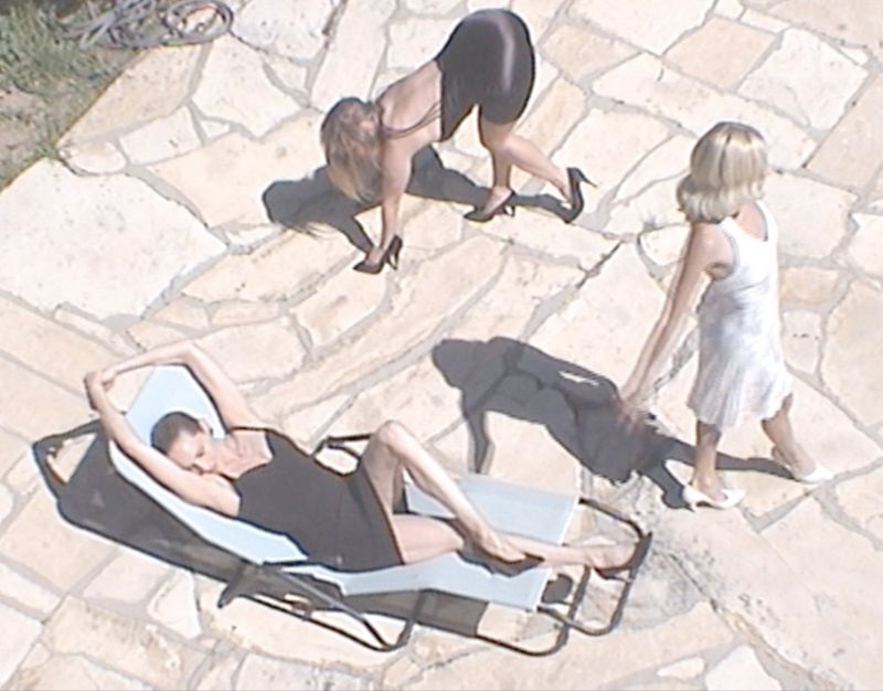 Overhead view of three adult women in dresses and heels posing outdoors on a stone patio in sunlight, with one woman reclining on a lounge chair.