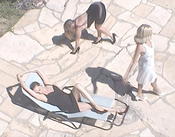 Overhead view of three adult women in dresses and heels posing outdoors on a stone patio in sunlight, with one woman reclining on a lounge chair.