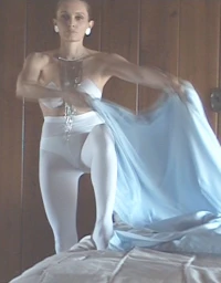 Woman in white tights and a bandeau top holding light blue draped fabric against a wood paneled wall.