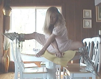 Blonde woman in a pale pink lace dress and patterned boots stretching across a white dining table in a wood-paneled room.