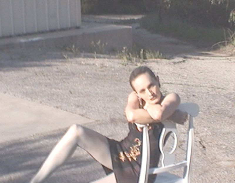 Model in a sheer black embroidered mini dress leaning over the back of a white chair outdoors on a paved lot.