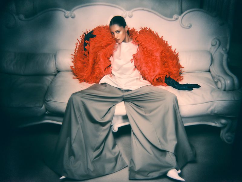 Model seated on a white sofa wearing a vibrant orange feathered jacket and wide grey trousers for Vogue Arabia.