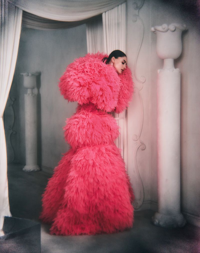 Model wearing a voluminous hot pink feathered gown standing in an architectural room with white columns.