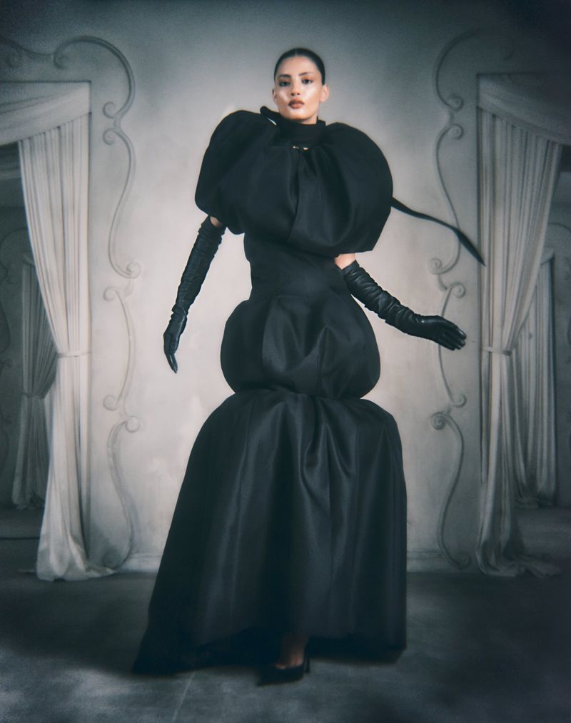 Model in voluminous black couture gown and long gloves standing before draped archways