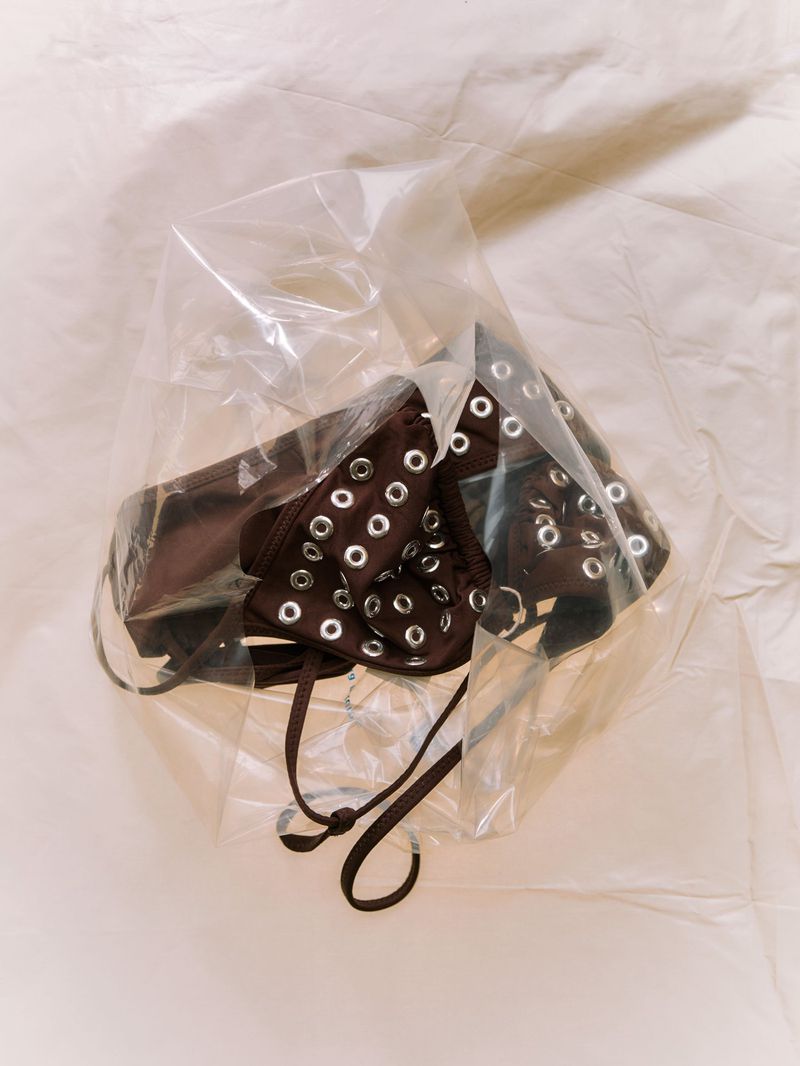 Brown garment with silver grommets enclosed in a crinkled translucent plastic bag on a neutral fabric background.