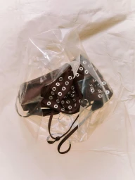 Brown garment with silver grommets enclosed in a crinkled translucent plastic bag on a neutral fabric background.