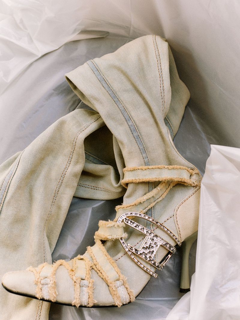 High heel denim boot with rhinestone buckle and distressed straps nestled inside white packaging paper