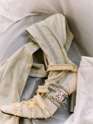 High heel denim boot with rhinestone buckle and distressed straps nestled inside white packaging paper