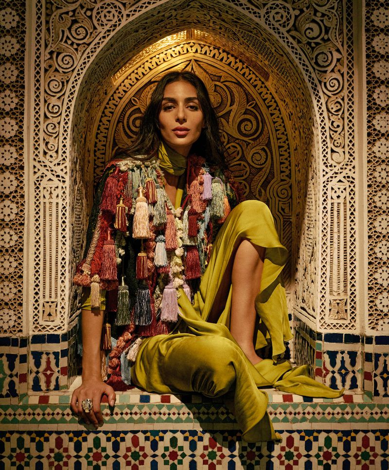 Model Nora Attal poses in an ornate arched alcove wearing a heavily tasseled garment and olive green silk trousers.