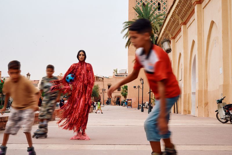 Model Nora Attal in red fringed dress holding blue football on public square with children roller skating