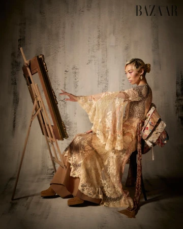 Model Lauren Nguyen in patterned gown seated beside an artist easel for Harper's Bazaar Vietnam
