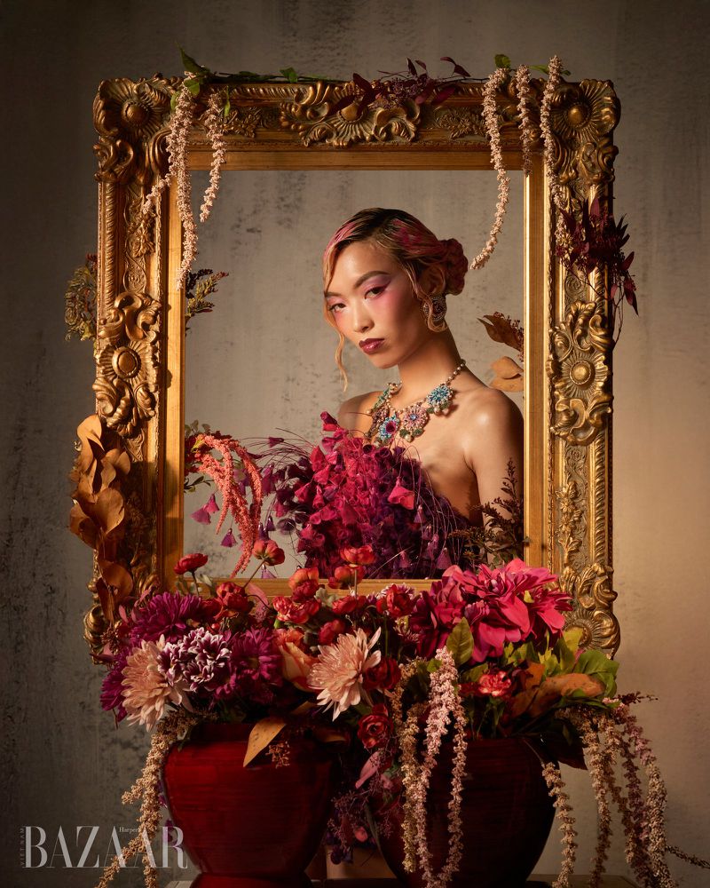 Model Lauren Nguyen posed behind an ornate gold picture frame adorned with floral arrangements and draped hanging plants.