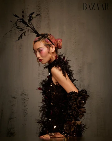 Model Lauren Nguyen with pink hair and feathered headpiece wearing a black feathered dress against a textured grey background.