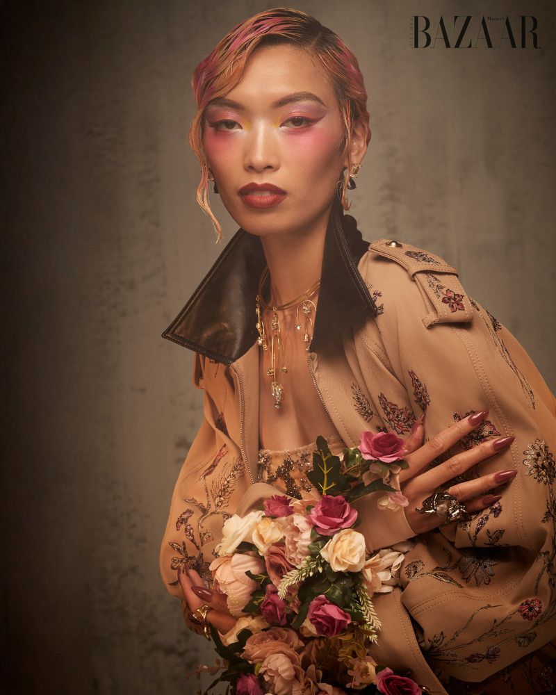 Model Lauren Nguyen holding a floral bouquet, wearing a beige embroidered jacket against a textured grey background.