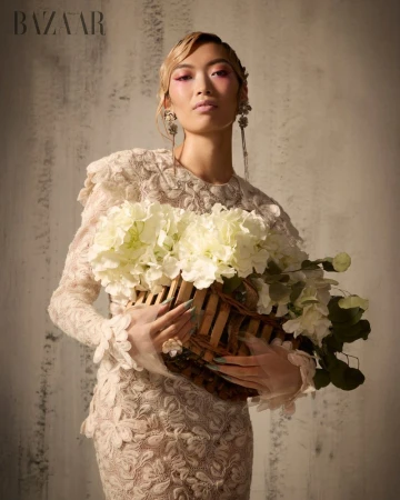 Model holding a basket of white hydrangeas wearing an off-white lace dress against a textured neutral wall