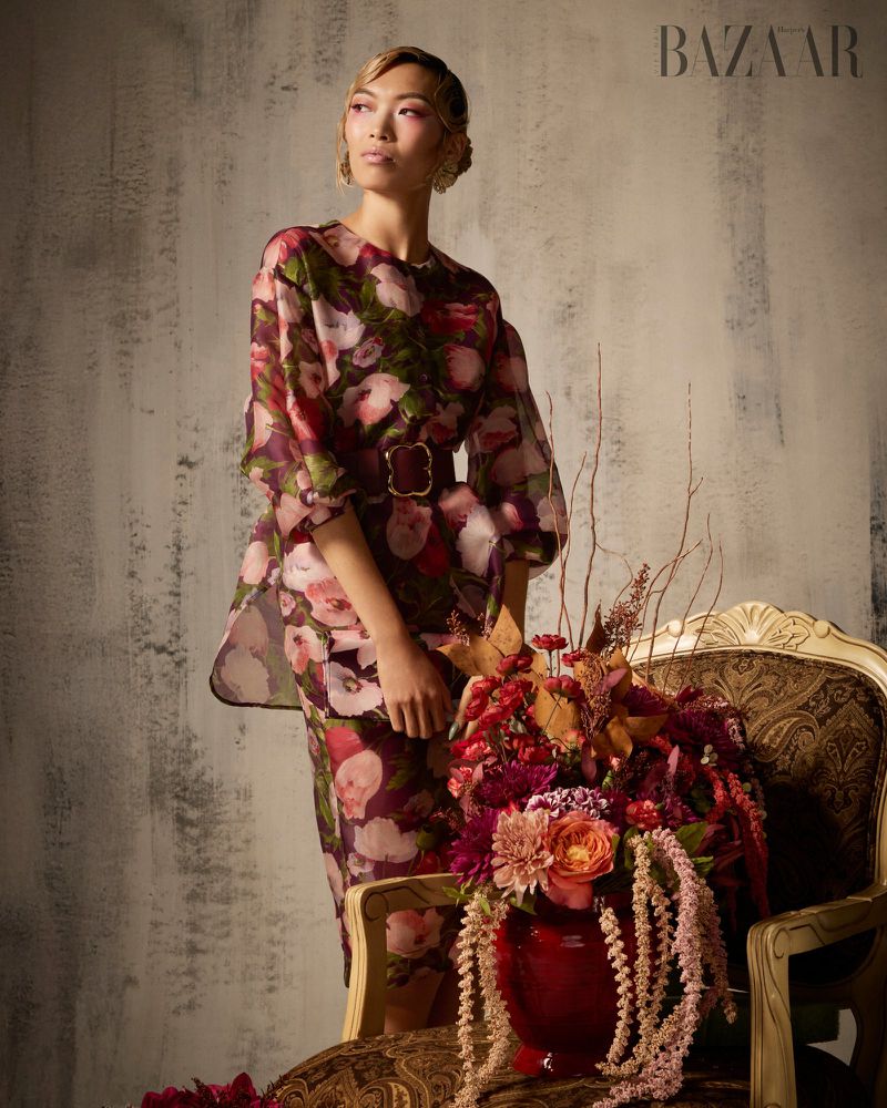 Model Lauren Nguyen wearing a floral sheer dress standing beside an ornate armchair with a lush floral arrangement.